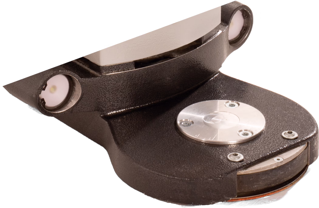 PALLMANN_dummy_Gecko Star attachment length short_msl_2018-07 Primer plano de una pieza mecánica robusta de metal fundido de color marrón oscuro/negro con acabado texturizado. La pieza tiene una base curva y una brida inclinada hacia arriba; en los bordes superiores izquierdo y derecho se aprecian dos pequeños elementos circulares empotrados que parecen luces o sensores. En la superficie superior plana hay instalada una tapa metálica redonda pulida con tres tornillos visibles, rodeada de varios tornillos avellanados. En el borde frontal se ve parcialmente un disco circular fino y giratorio o un componente parecido a un freno con un borde naranja. La imagen está recortada sobre fondo blanco y muestra la textura detallada de la superficie, las cabezas de los tornillos y los reflejos metálicos.