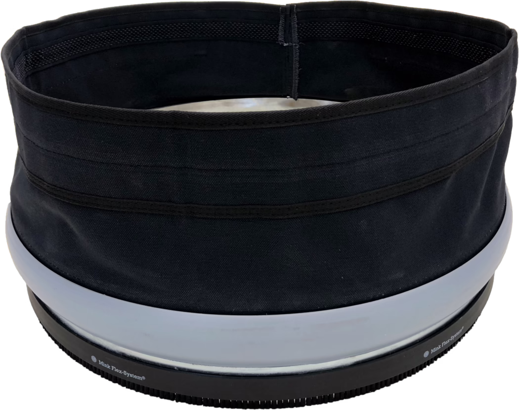Primer plano del borde superior y la apertura de un contenedor cilíndrico de tela negra o un sombrero. La imagen muestra una abertura circular interior con forro negro, costuras reforzadas visibles y varias bandas horizontales de tela en el exterior. Cerca del borde inferior hay dos anillos anchos de plástico gris claro y una base de goma negra acanalada con pequeños dientes y texto impreso parcialmente visible 'Mink Flex-System®'. La tela tiene un tejido toscamente entrelazado y un acabado mate; el objeto parece estructurado pero flexible. La iluminación es uniforme y el fondo es blanco.