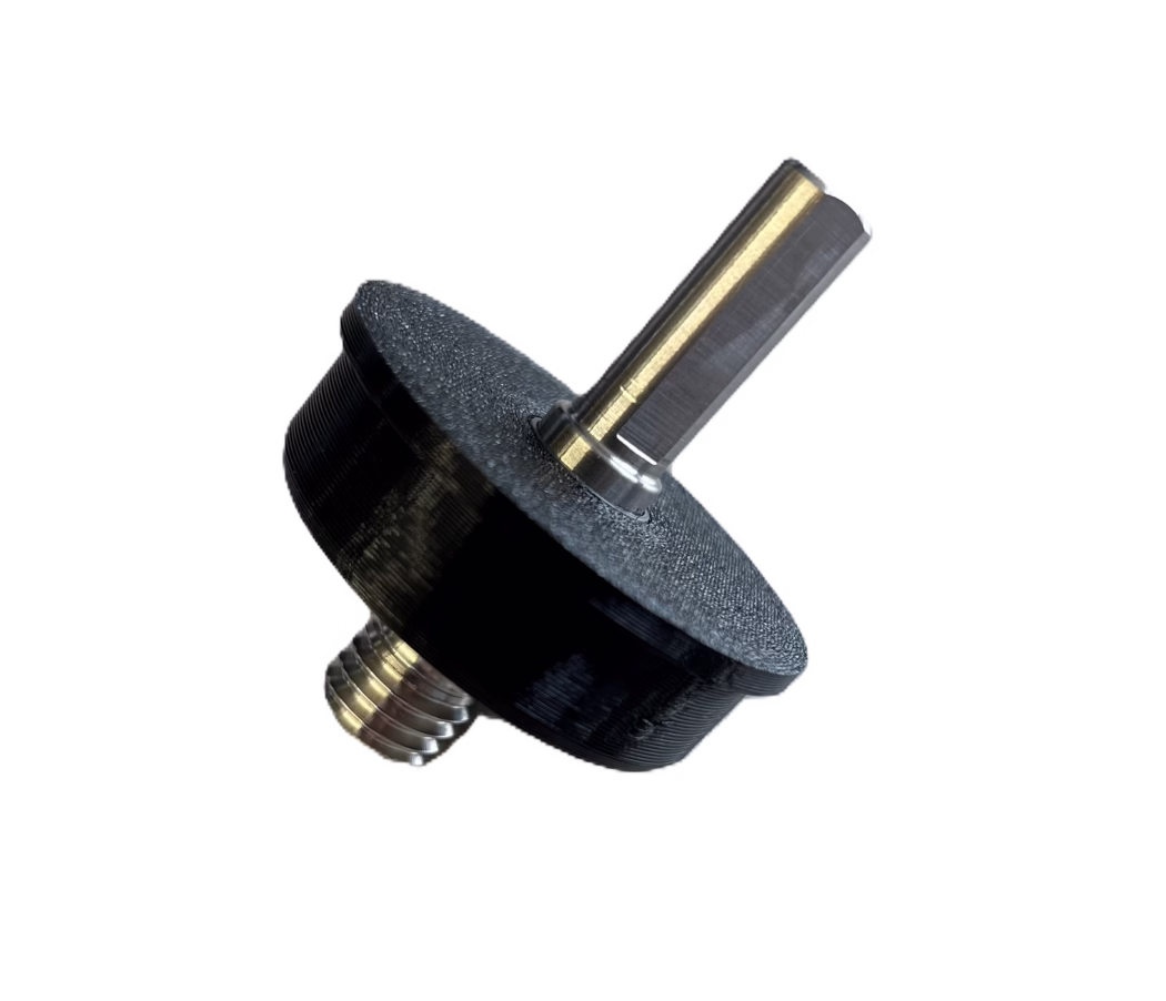 PALLMANN_product image_adapter cordless screwdriver Plano detalle de un pequeño conjunto mecánico: un eje metálico hexagonal con recubrimiento color latón insertado en una pieza negra de plástico o en un mando/espaciador impreso en 3D. El eje metálico muestra una cara plana pulida y marcas de mecanizado visibles; en la base se ve un vástago roscado con hilos concéntricos. La pieza negra tiene una superficie texturizada y estratificada, típica de impresión 3D FDM. El objeto está fotografiado en diagonal sobre un fondo blanco limpio.